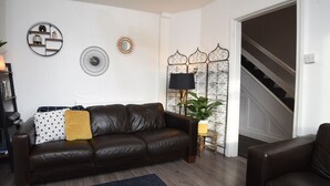 A living room with a brown leather sofa, a small side table with a lamp, a potted plant, and a staircase leading to another level. - Cosy and Modern 2 Bed Family House in Abergele (Abergele)