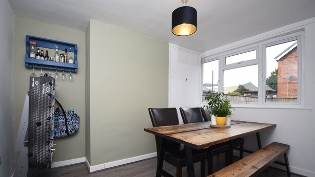 A dining area with a wooden table, black chairs, and a bench. There is a hanging rack with bags and bottles, a potted plant on the table, and a window with a view of a brick building. - Cosy and Modern 2 Bed Family House in Abergele (Abergele)