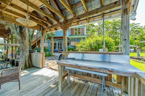 Une terrasse en bois, agrémentée d’une table et de chaises, entourée d’une pergola rustique en bois, donnant sur un jardin avec une maison et de la verdure.