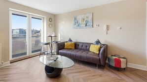 A modern living room with a glass coffee table, a brown leather sofa with cushions, a small stool, and a patchwork ottoman. - Properties Unique | The Peak (Newcastle Upon Tyne)