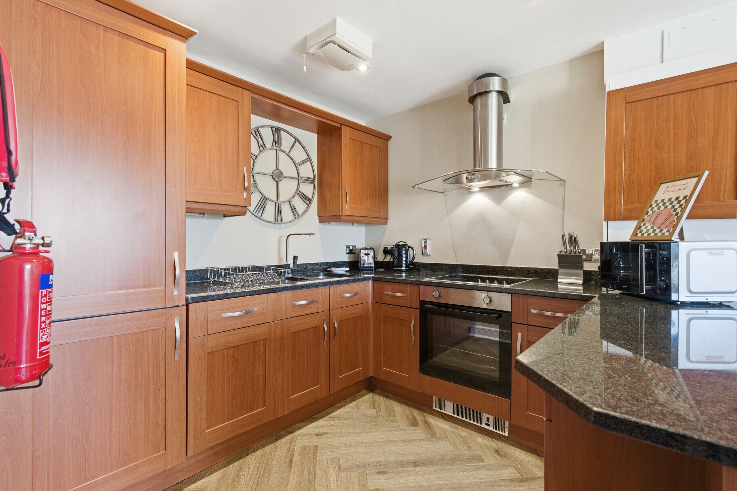 A kitchen with wooden cabinets, a stainless steel range hood, a clock, and a fire extinguisher.