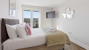 A bedroom with a bed, a TV, a window, and a wall-mounted mirror. - Properties Unique | The Peak (Newcastle Upon Tyne)