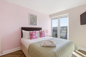 Un dormitorio con cama, almohadas, un cuadro, una mesita de noche y una ventana.