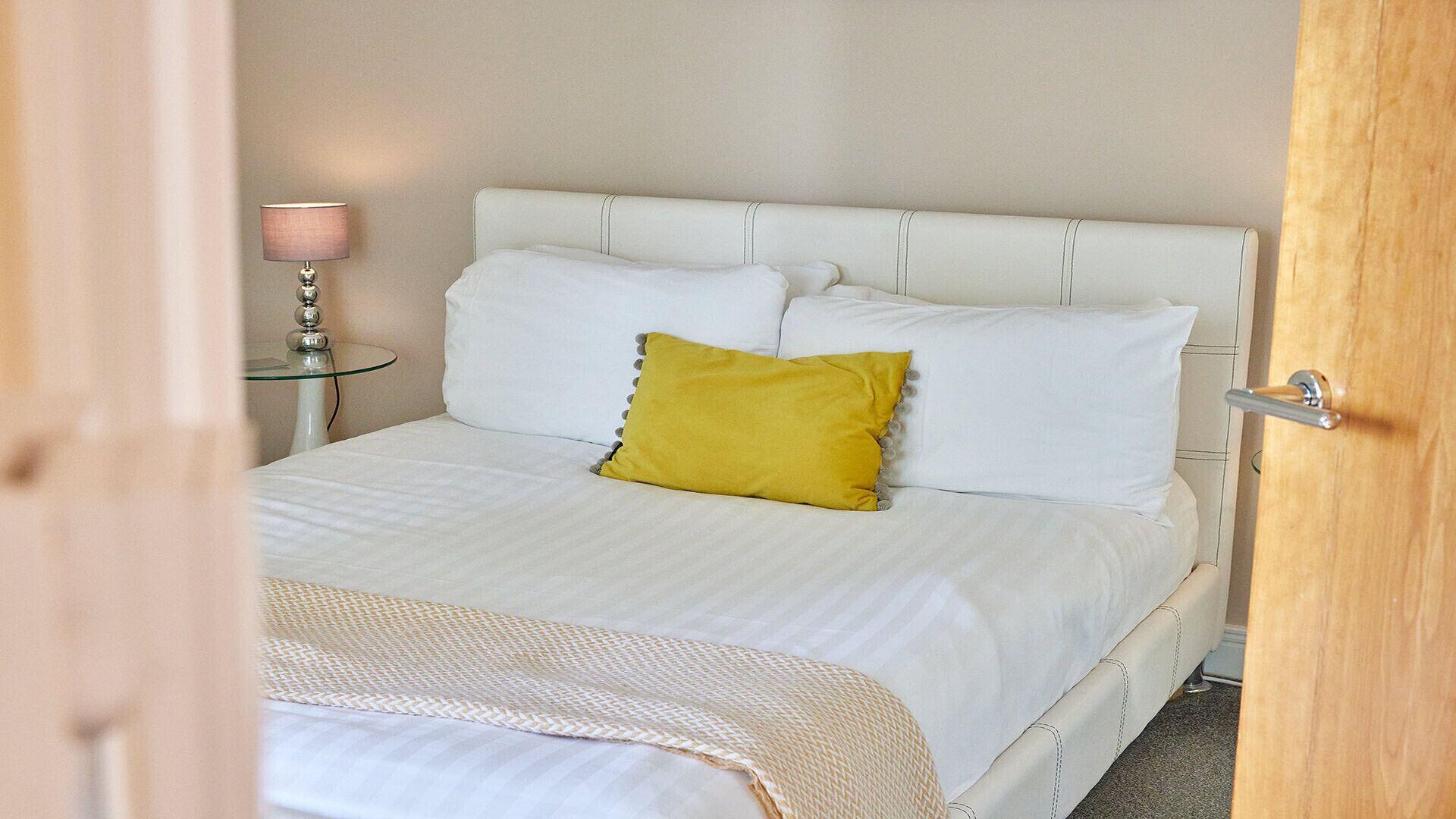 Un letto con biancheria bianca e un solo cuscino giallo, un comodino con una lampada e una porta in legno.