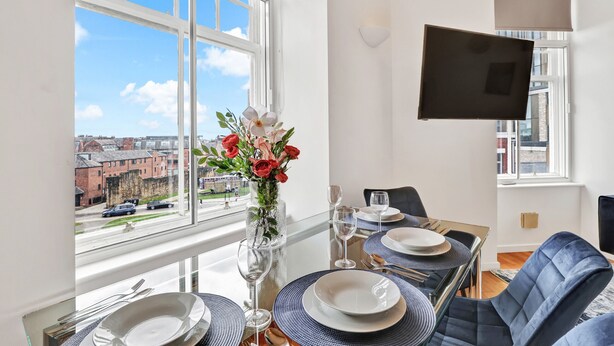 A dining area with a table set for four, a vase of flowers, and a large window with a city view. - Properties Unique | The Loft (Newcastle Upon Tyne)