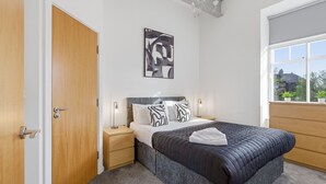 A bedroom with a bed, bedside tables, a wall-mounted artwork, a window with a view, and a door. - Properties Unique | The Loft (Newcastle Upon Tyne)