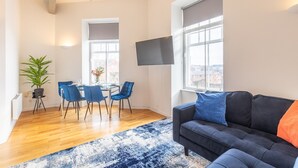 A modern living room with a sofa, a dining table with chairs, a TV mounted on the wall, and large windows. - Properties Unique | The Loft (Newcastle Upon Tyne)