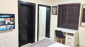 A bedroom with a bed, a desk, a chair, a TV displaying movie posters, and a window with blinds. - The Leneade - Exquisite Apt.1 (City Centre/Wuse 2) (Abuja)
