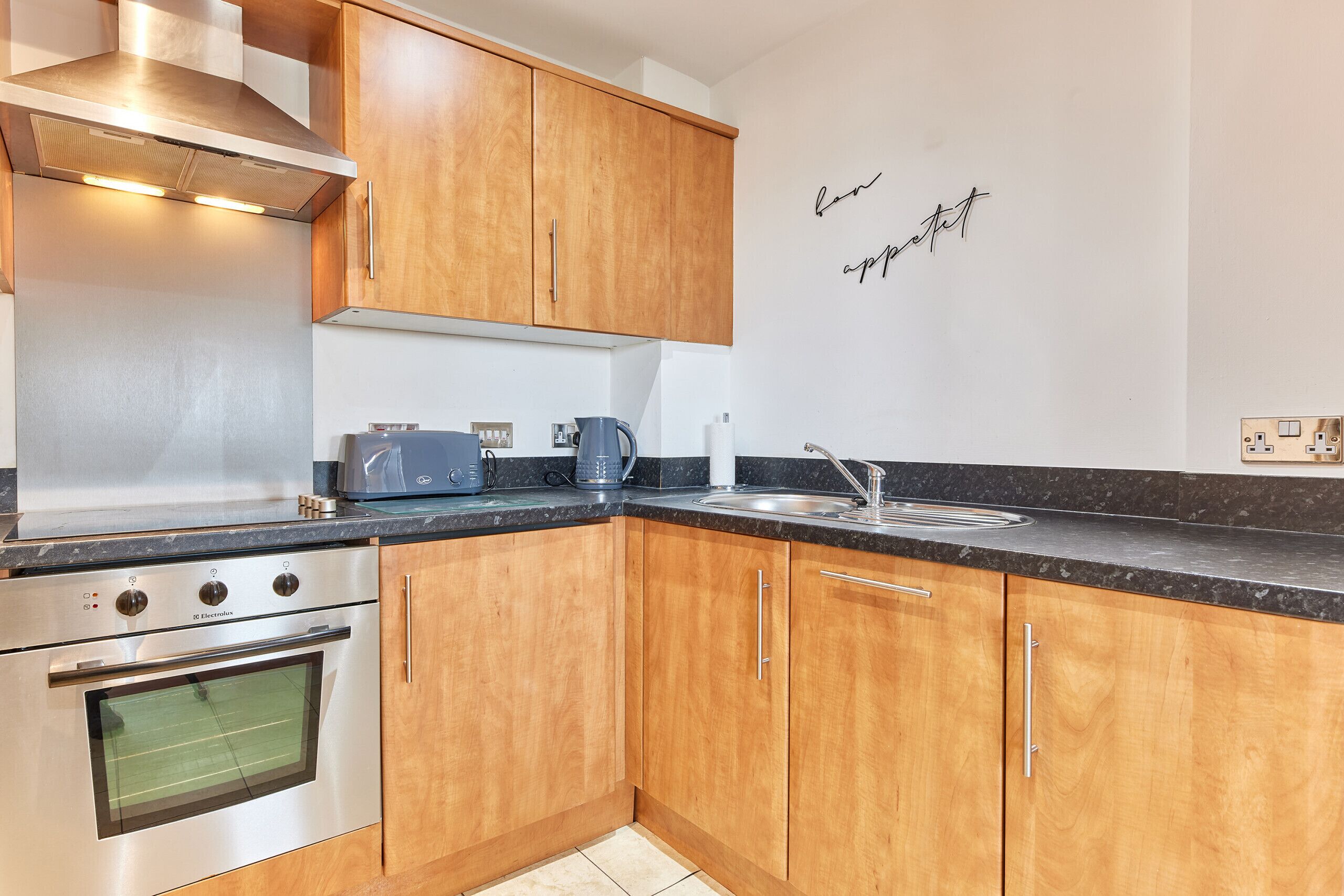 A modern kitchen with wooden cabinets, stainless steel appliances, a built-in oven, a coffee maker, and a wall-mounted light switch.