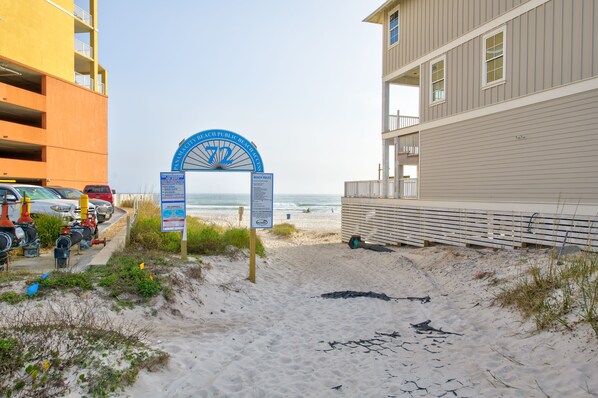 A beach entrance with a sign indicating beach rules, a parking area with cars, and a building with balconies overlooking the beach. - STEPS TO THE BEACH ❤️Romantic! (Panama City Beach)