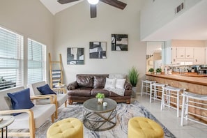 A modern living room with a brown leather sofa, a round coffee table, and yellow ottomans. There is a kitchen area with white cabinets and a bar counter. - STEPS TO THE BEACH ❤️Romantic! (Panama City Beach)