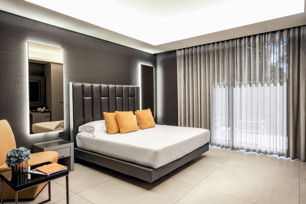 Ein modernes Hotelzimmer mit einem großen Bett, einem Nachttisch, einem Sessel, einem Spiegel und einem großen Fenster mit Vorhängen.