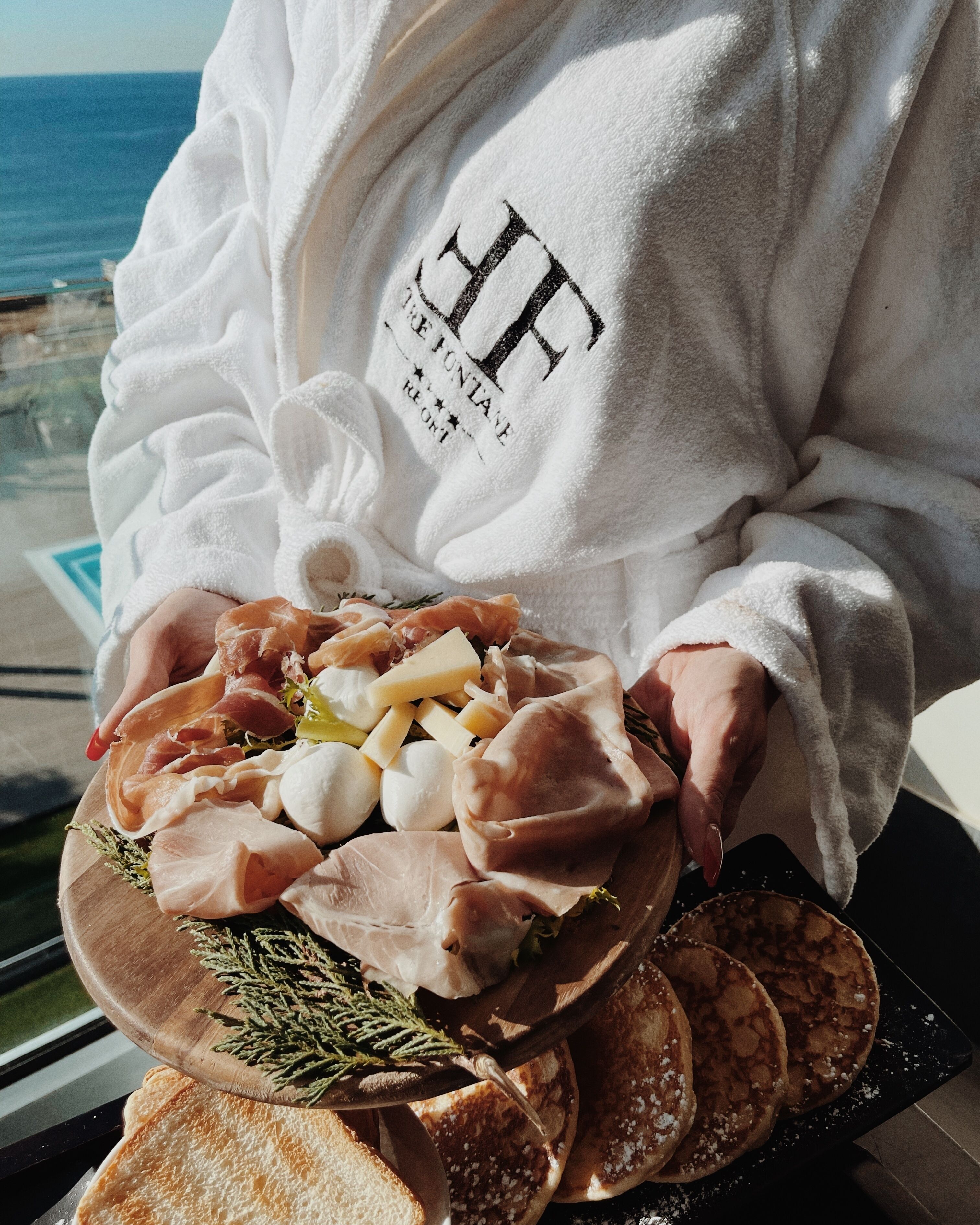 A person holding a charcuterie board with various meats, cheese, and bread, with a view of the ocean in the background.