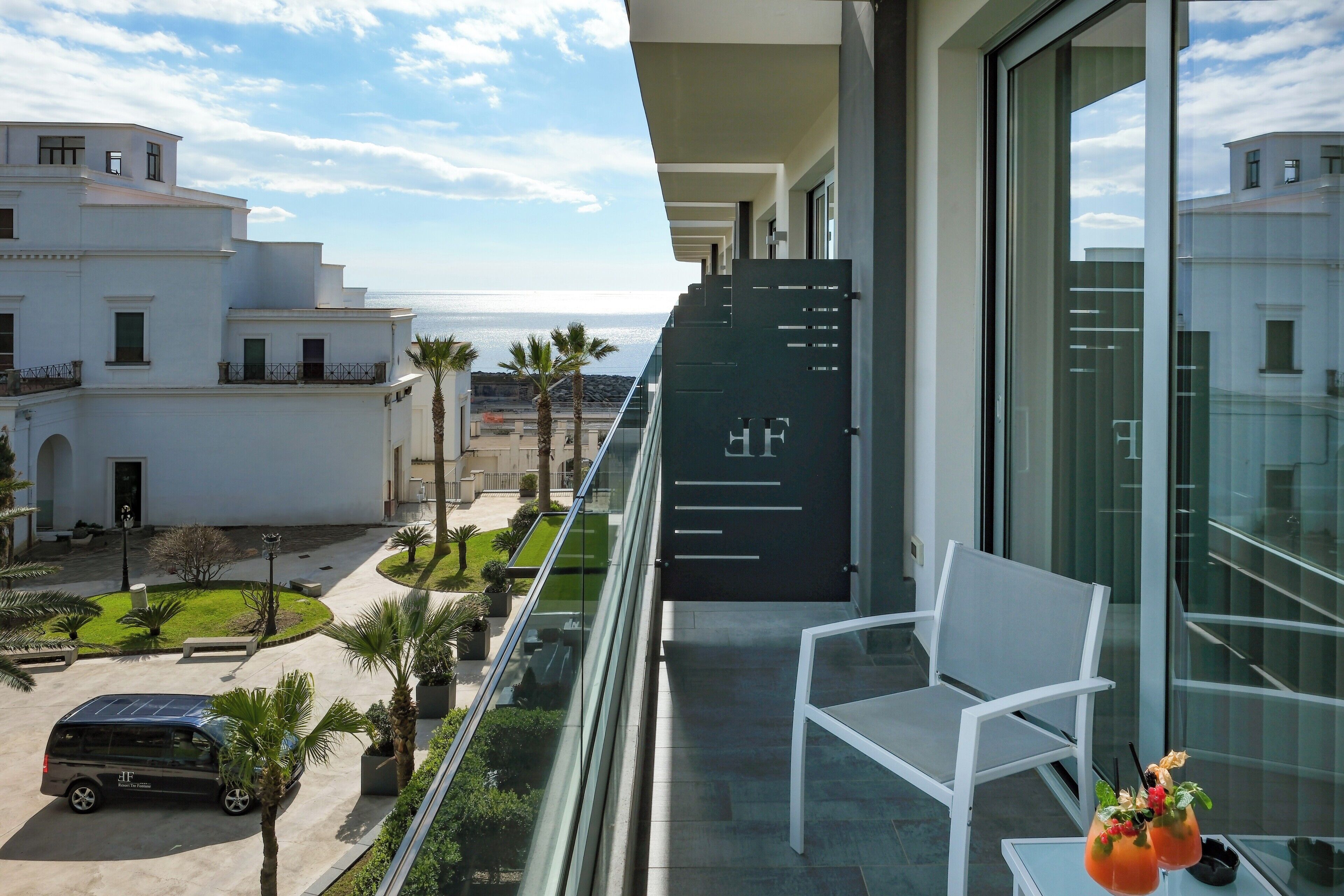 Un balcón con una silla blanca, una mesa con bebidas y vistas al océano y edificios.
