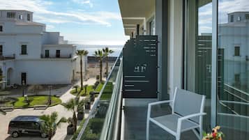 Un balcón con una silla blanca, una mesa con bebidas y vistas al océano y edificios.