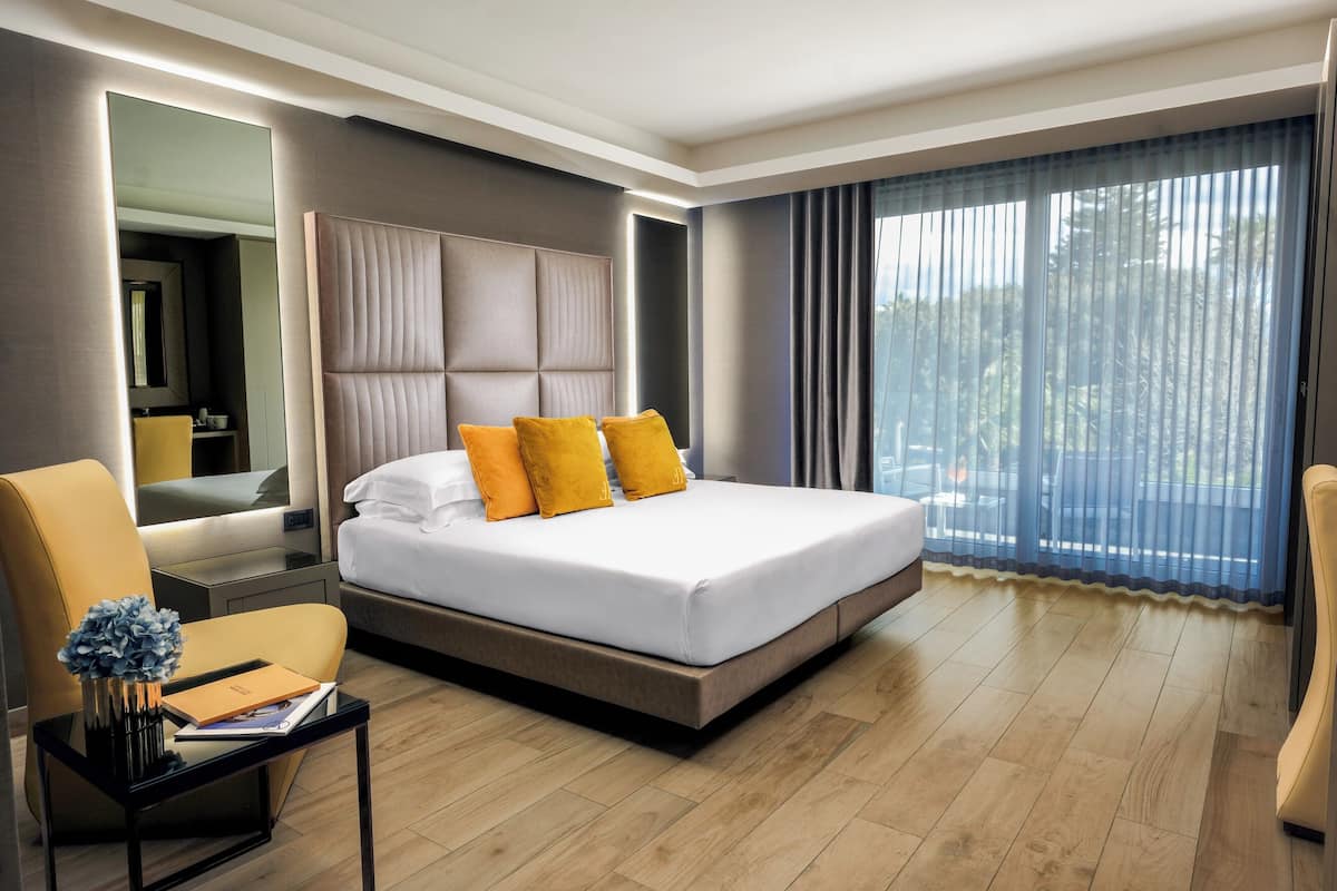 Ein modernes Hotelzimmer mit einem großen Bett, einem Nachttisch mit Buch und Vase, einem Sessel und einem großen Fenster mit Vorhängen.