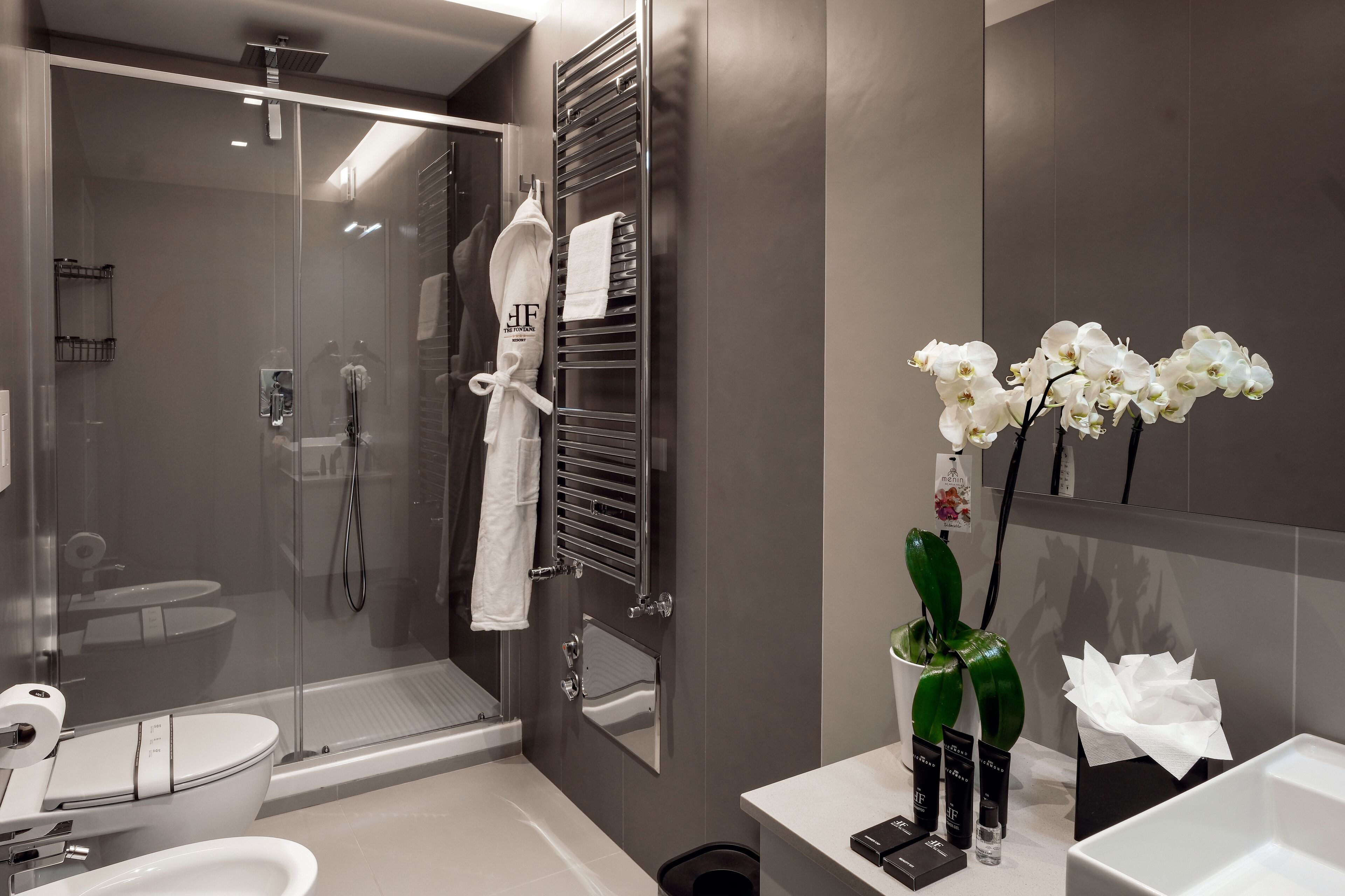 Een moderne badkamer met een douchecabine van glas, een wit toilet, een wastafel met toiletartikelen en een spiegel met een vaas witte orchideeën.