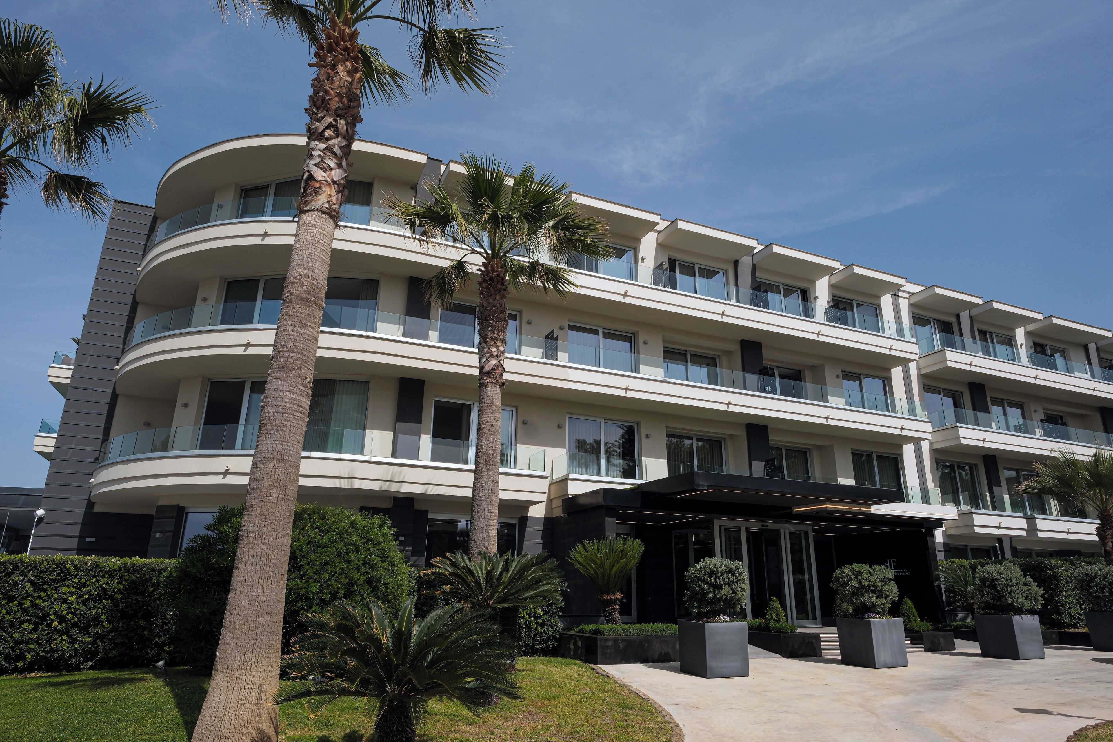 A multi-story hotel with a curved facade, palm trees, and a well-maintained garden.