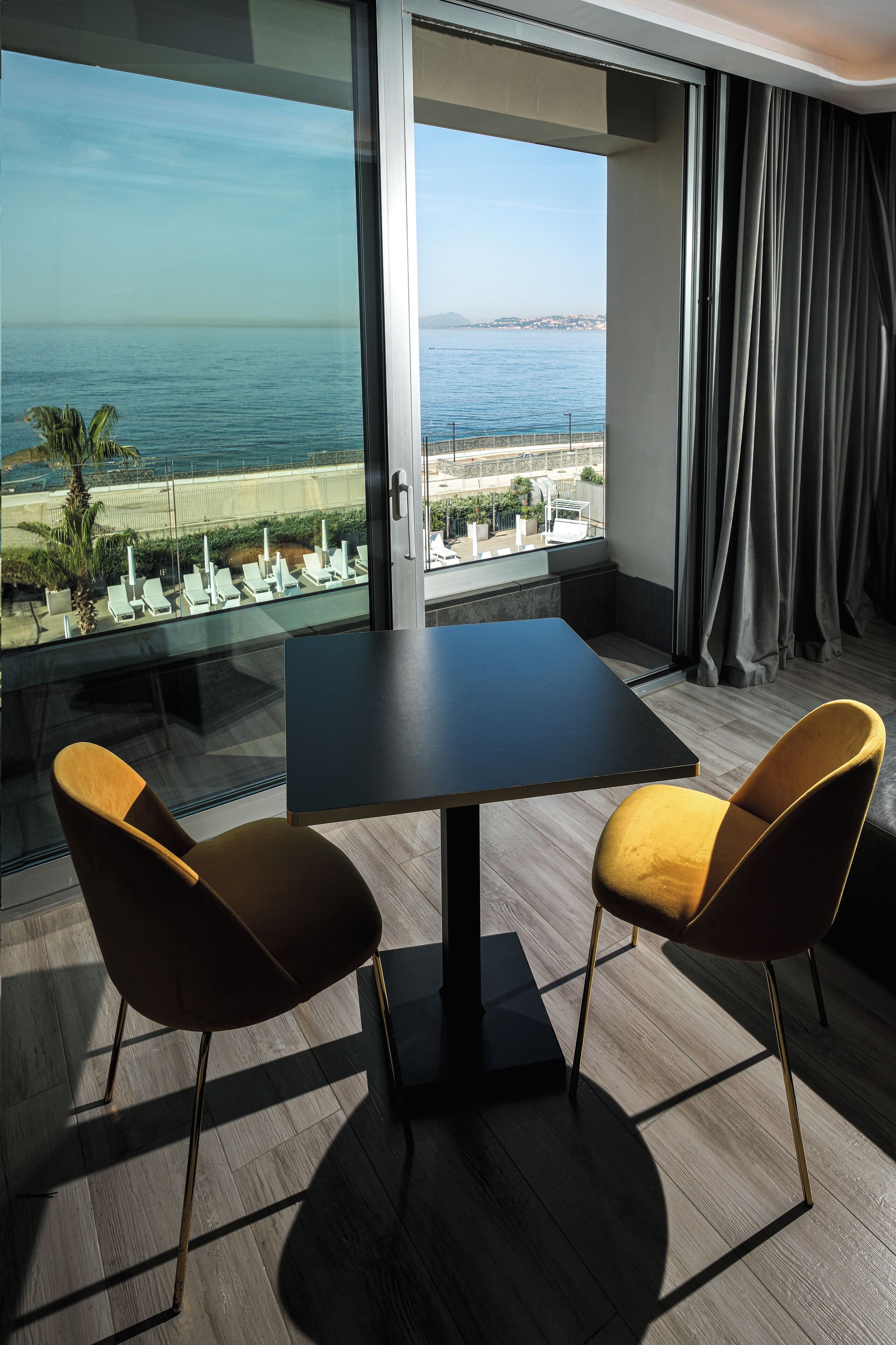 A room with a large window overlooking a beach, a dark table, and two yellow chairs.