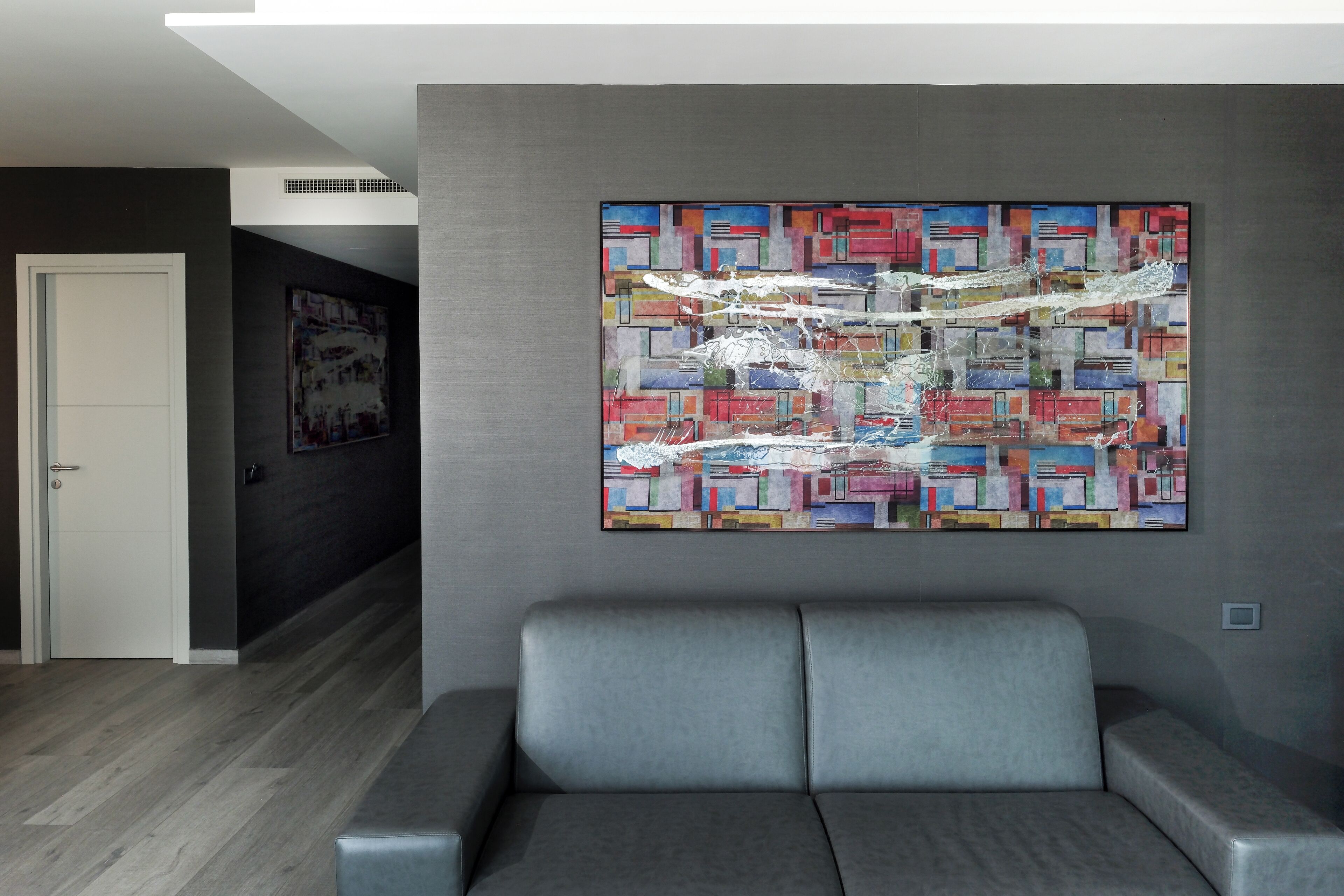 Ein moderner Wartebereich mit einem grauen Sofa, einem farbenfrohen abstrakten Gemälde an der Wand und einer Tür im Hintergrund.