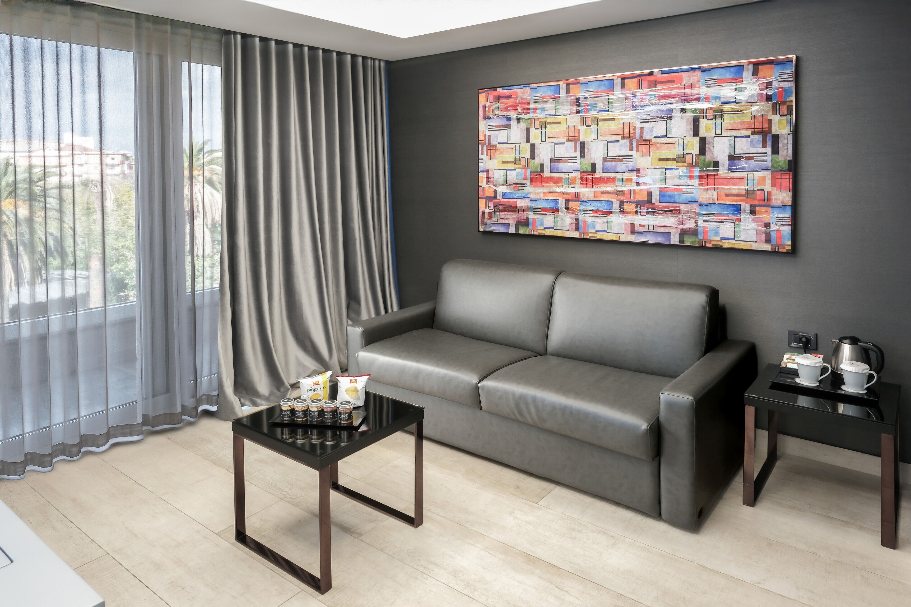 A modern living room with a grey leather sofa, a black coffee table, and a small side table with a kettle and cups. A colorful abstract painting is on the wall, and a window with sheer curtains is visible.