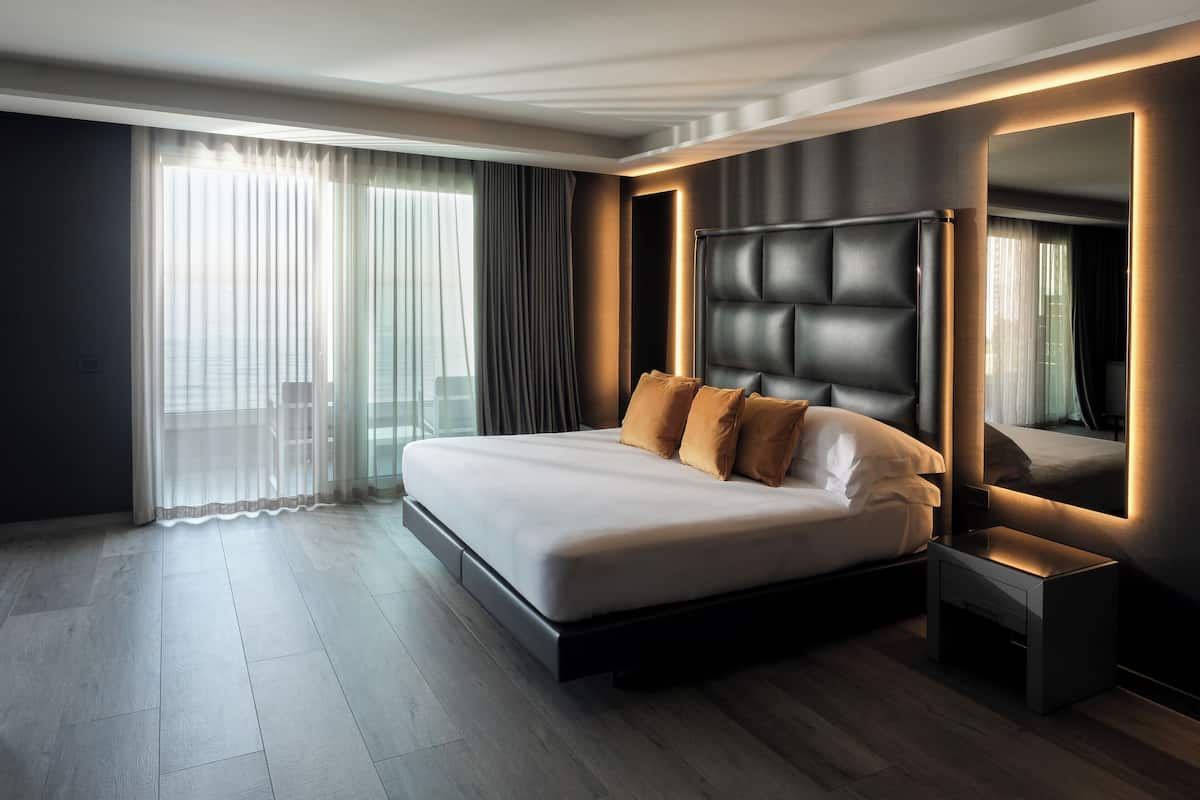 Ein modernes Hotelzimmer mit einem großen Bett, einem Nachttisch, einem Spiegel und Blick auf die Stadt durch große Fenster.
