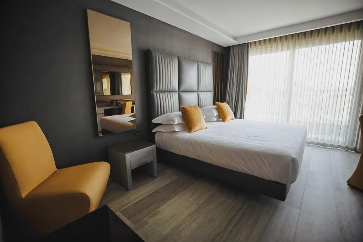 A modern hotel room with a large bed, a chair, a nightstand, and a mirror.