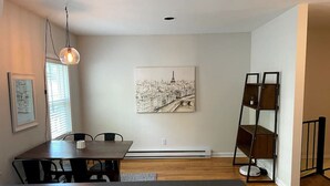 A modern living room with a dining area, a large painting of a cityscape, a standing heater, and a black metal shelf unit.