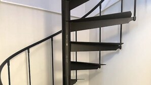 A spiral staircase with a black metal railing, white walls, and brown tiled flooring.