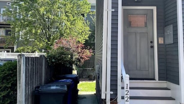 A residential house with a gray exterior, a white front door, and a small porch with steps. There are two trash bins outside the door.