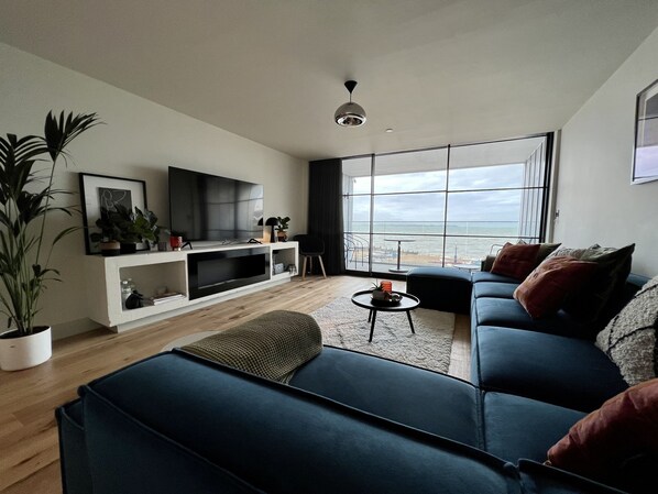 A modern living room with a large TV, a sofa, a coffee table, and a view of the sea through large windows.