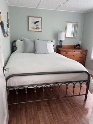 Ein Schlafzimmer mit einem Metallbettgestell, einer Holzkommode, einem Nachttisch mit Lampe und einem gerahmten Bild an der Wand.