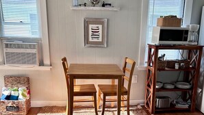 A dining area with a wooden table and chairs, a woven rug, and a shelf with kitchen items.