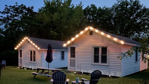 Two cabins with string lights, a picnic table, and chairs in a grassy area.