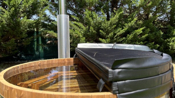 Un bain à remous en bois, doté d’une cheminée en métal, entouré d’arbres.