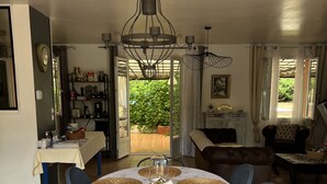 Un coin repas avec une table ronde dressée pour six personnes, entourée de chaises bleues, d’un lustre et offrant une vue sur un jardin par une porte ouverte.