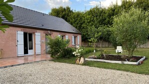 Une maison avec un toit en tuiles, une allée gravillonnée et un jardin où poussent différentes plantes ainsi qu’un petit arbre.
