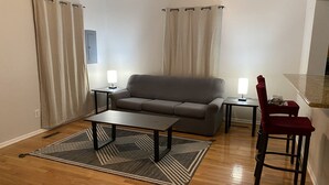 Een woonkamer met een bank, salontafel, twee bijzettafels met lampen, een rode fauteuil, en een keuken met een aanrecht.