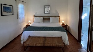 A bedroom with a bed, bedside lamps, a framed picture on the wall, a window with curtains, and a view of the outside. - Pousada Villa Cumuru (Prado)