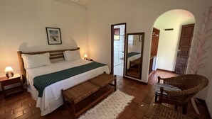A bedroom with a bed, bedside tables, a bench, a chair, a mirror, and a door leading to another room. - Pousada Villa Cumuru (Prado)