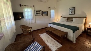A bedroom with a bed, a chair, a bench, a TV, and a window with curtains. - Pousada Villa Cumuru (Prado)