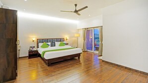 A hotel room with a wooden floor, a bed with green and white bedding, a ceiling fan, a nightstand with a lamp, and a view of the outdoors through a glass door. - Treebo Shindola Resort (Mahabaleshwar)