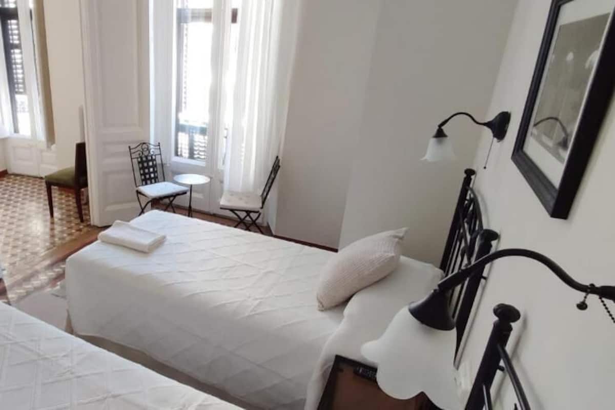 A hotel room with two beds, a chair, a small table, and a window with curtains.