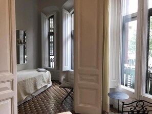 A bedroom with a bed, a small table, and a chair. There are windows with shutters and a view of the outside. - Casa Consell Bailen, Guest House  (Barcelona)