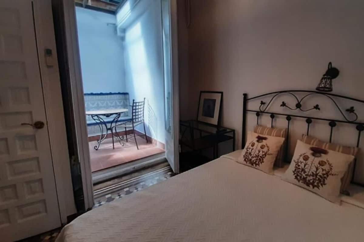 A bedroom with a bed, a nightstand, and a chair. There is a door leading to a balcony with a table and chairs.