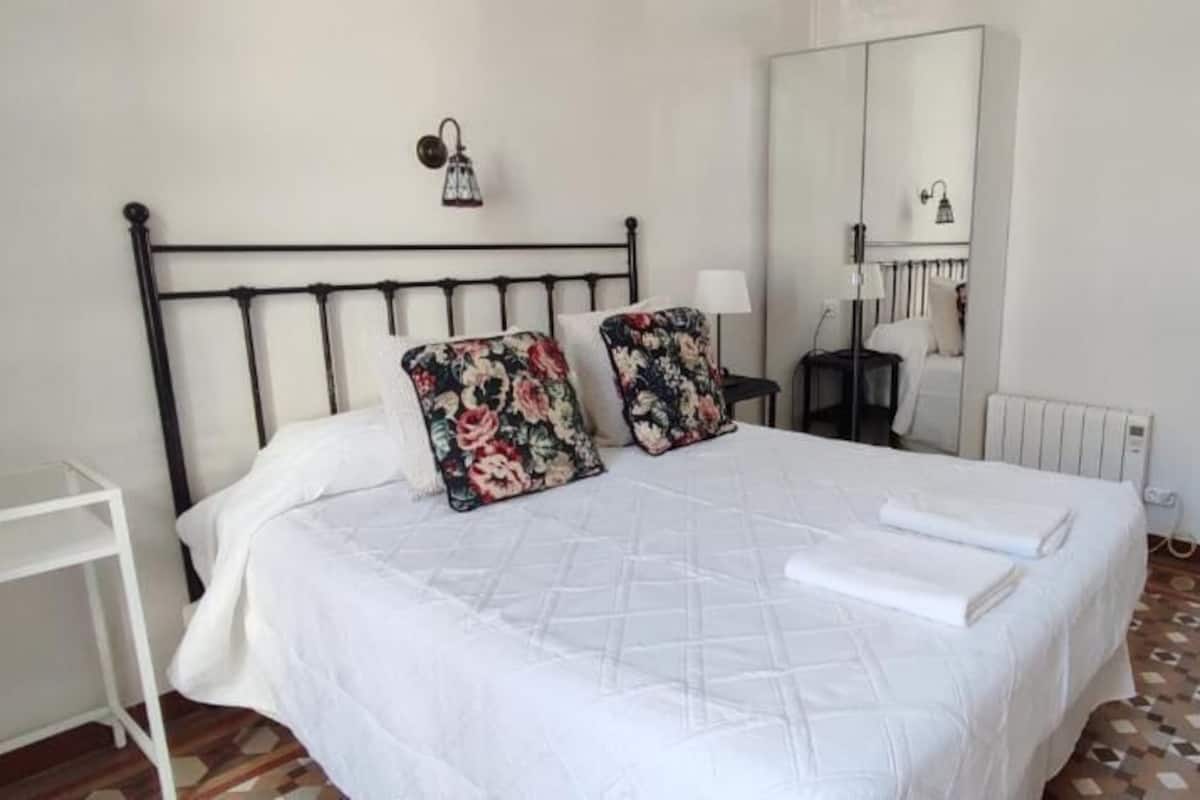 A bedroom with a bed, two floral pillows, a white duvet, a bedside table, a lamp, a mirror, and a radiator.