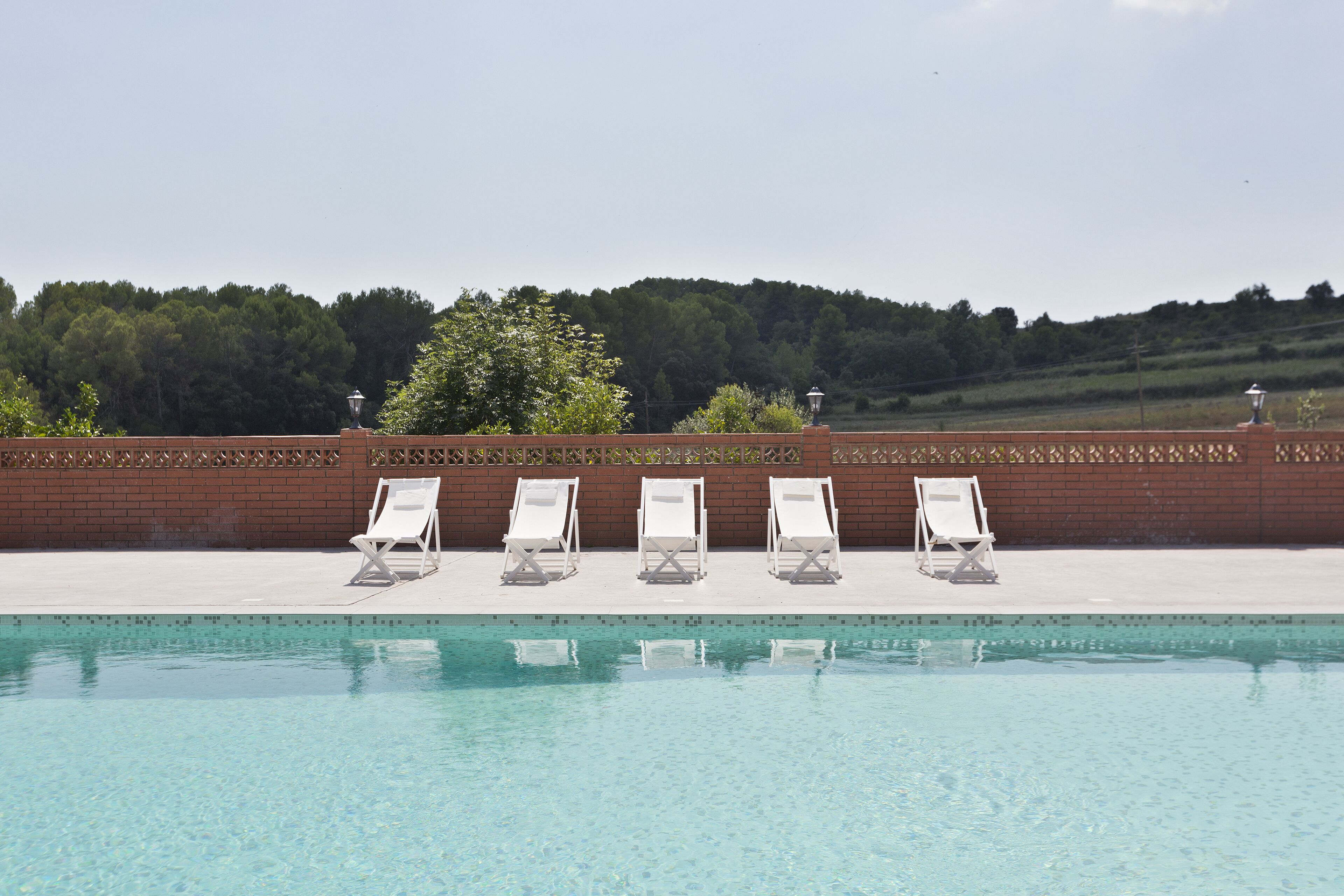 A swimming pool with clear water, surrounded by a brick wall and three white lounge chairs.