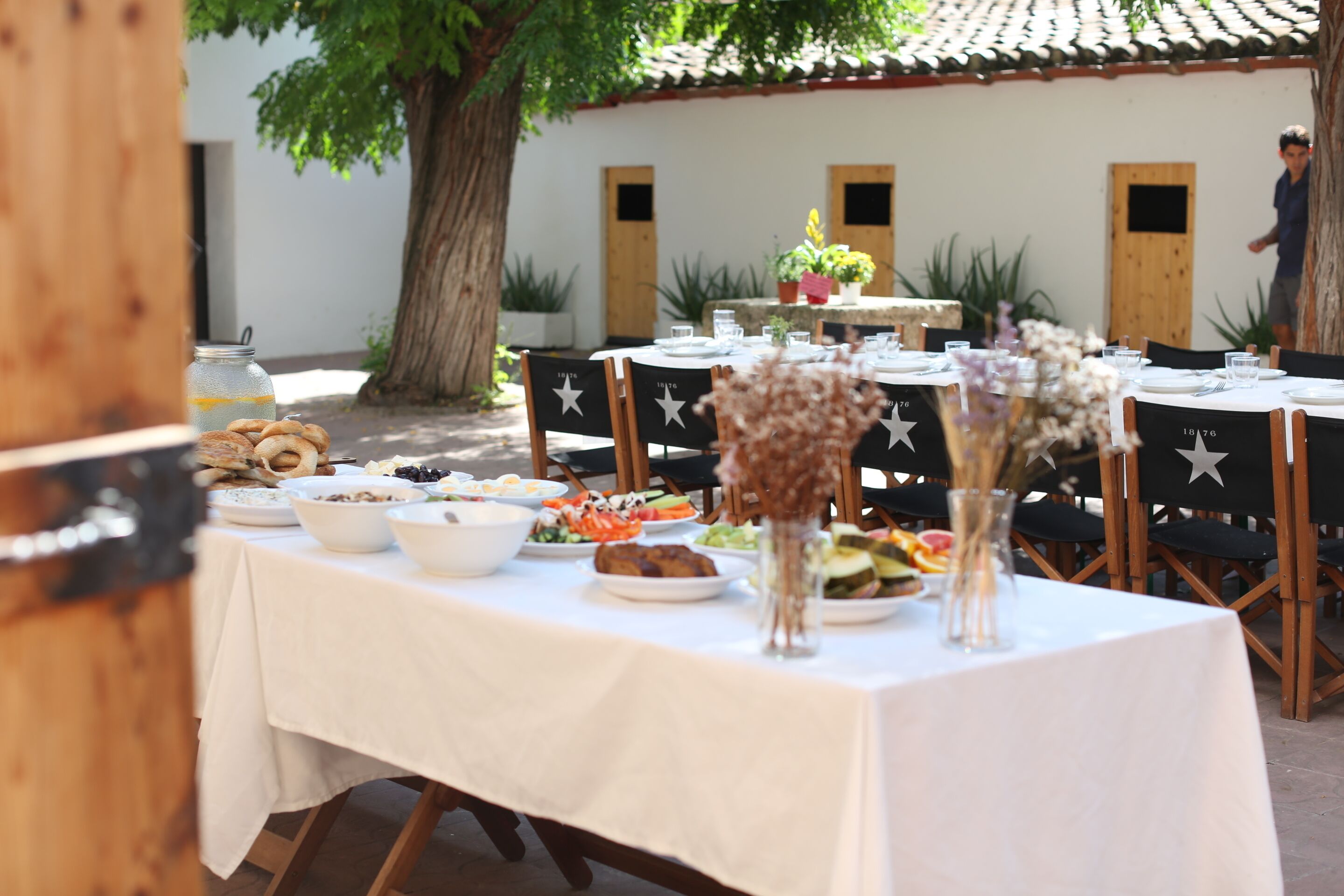 Un área de comedor al aire libre con mesas preparadas para una comida, rodeada de plantas en macetas y un árbol.
