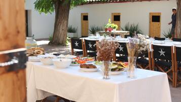 Un área de comedor al aire libre con mesas preparadas para una comida, rodeada de plantas en macetas y un árbol.