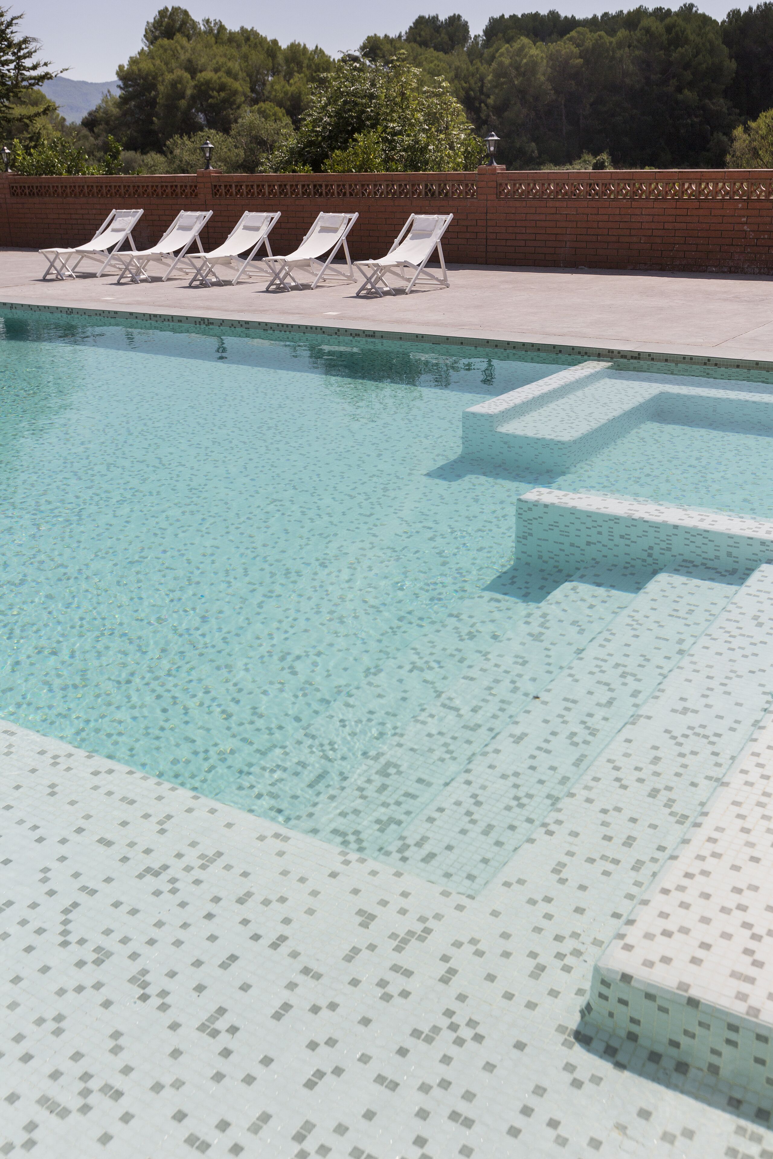 Une piscine à l’eau claire, entourée d’une terrasse carrelée et d’un mur en briques.